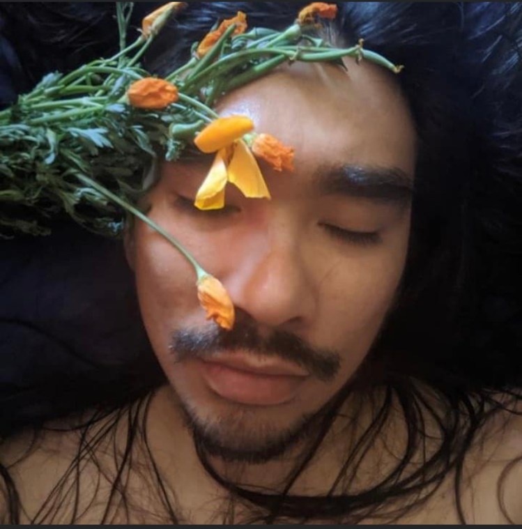 A picture of Edi Zermeno, a light brown skinned Mexican trans femme. She has dark brown long hair and a goatee and their eyes are closed. There are orange flowers strewn across her face. 